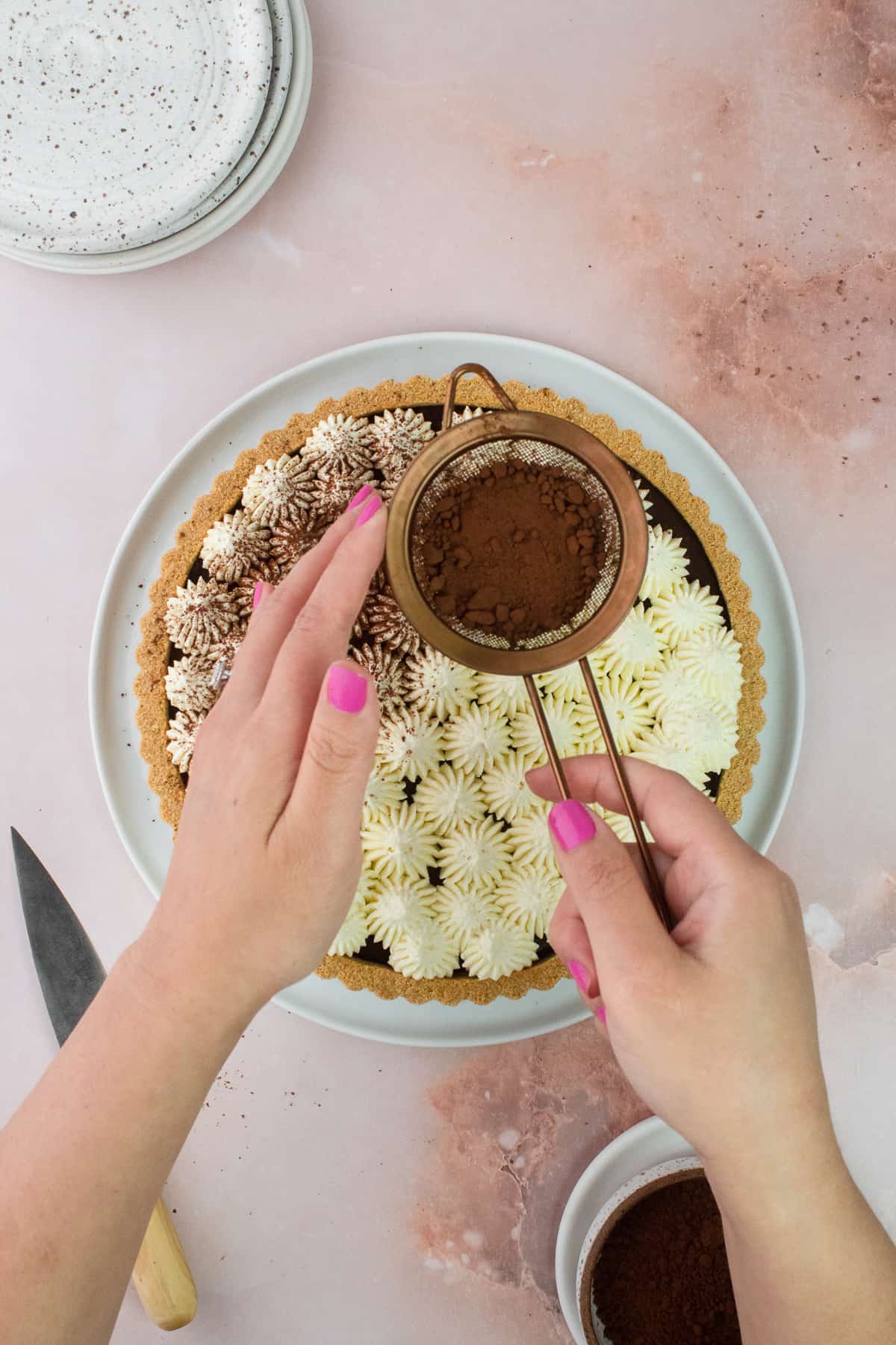 Hands are holding a small strainer with cocoa powder and dusting cocoa powder on top of the tart