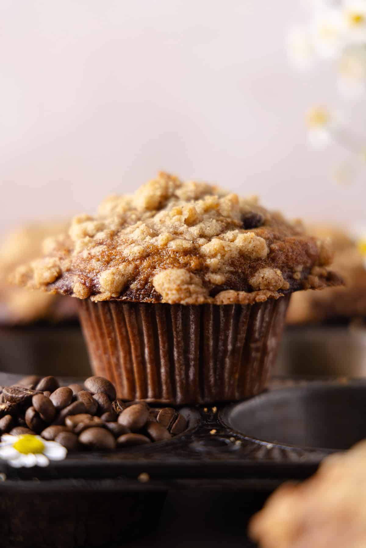 Close up of a Banana Espresso Muffin standing on top of a muffin tin with whole espresso beans in a cavity of the muffin tin.