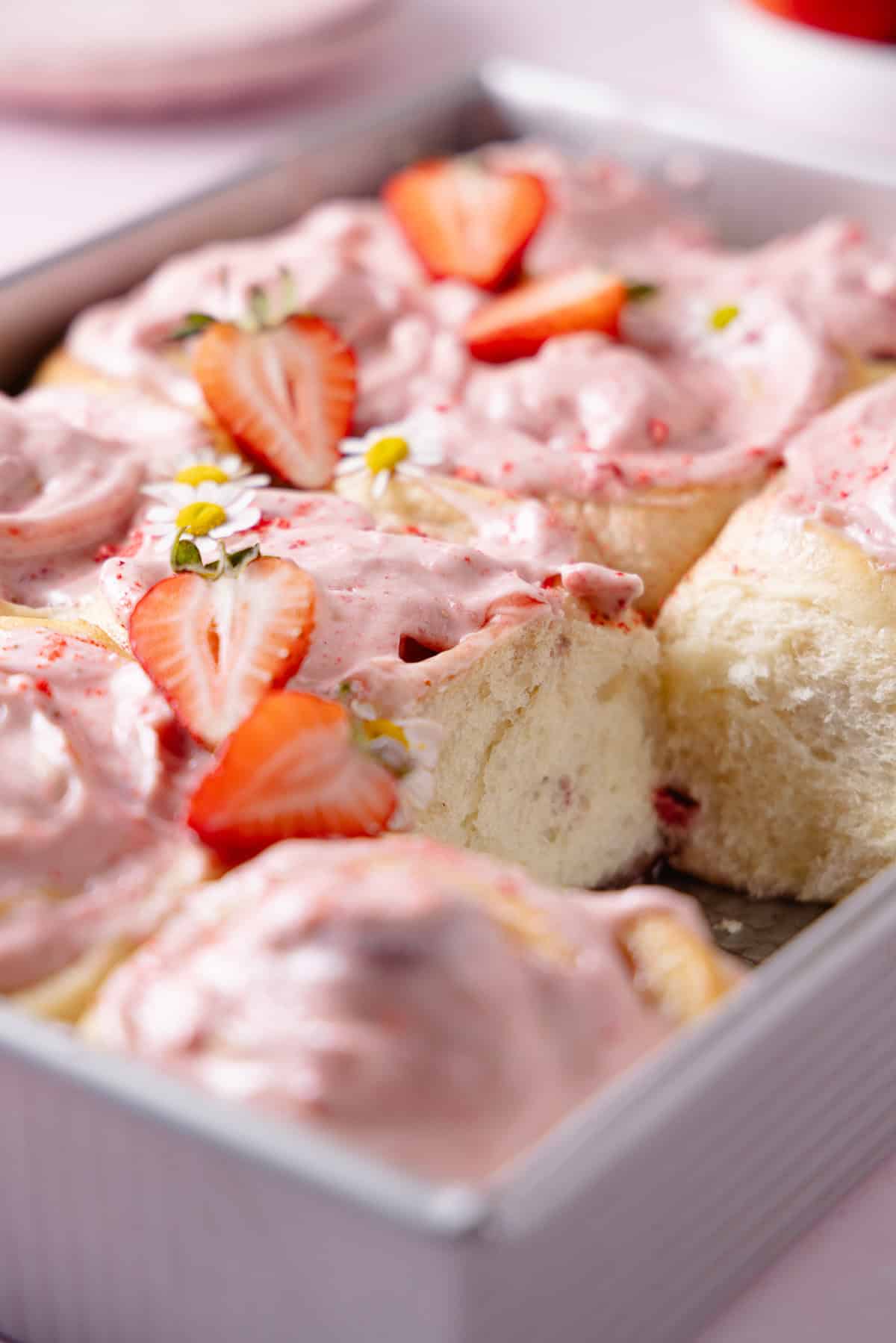 Frosted Strawberry Cinnamon Rolls in a baking tray garnished with fresh strawberries and flowers. A roll is missing from the tray, showing fluffy texture.
