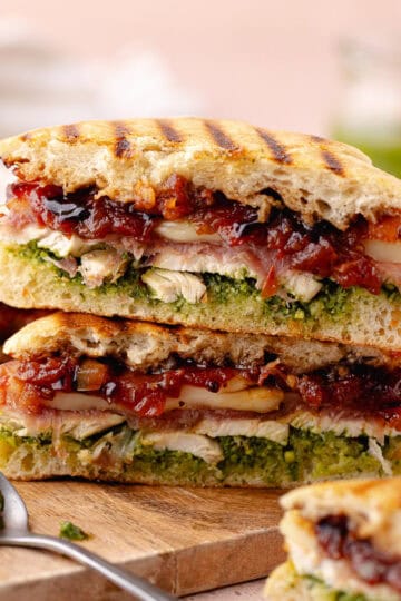 Chicken caprese panini (panino) filled with prosciutto, chicken breast, pistachio basil pesto, tomato jam, mozzarella cheese, balsamic glaze, cut in half and sitting on a small cutting board.