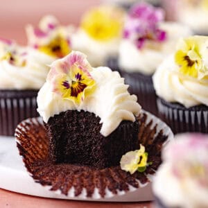 Chocolate cupcakes with cream cheese frosting decorated with edible flowers are sitting on a marble serving board feature photo.