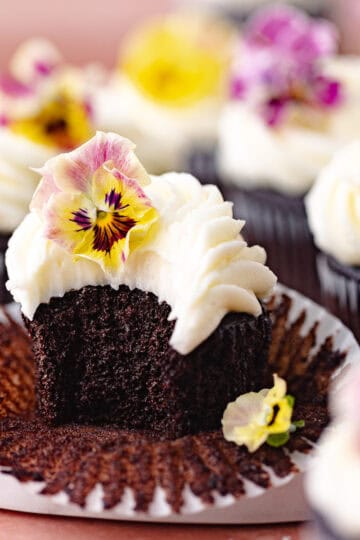 Chocolate cupcakes with cream cheese frosting decorated with edible flowers are sitting on a marble serving board feature photo.