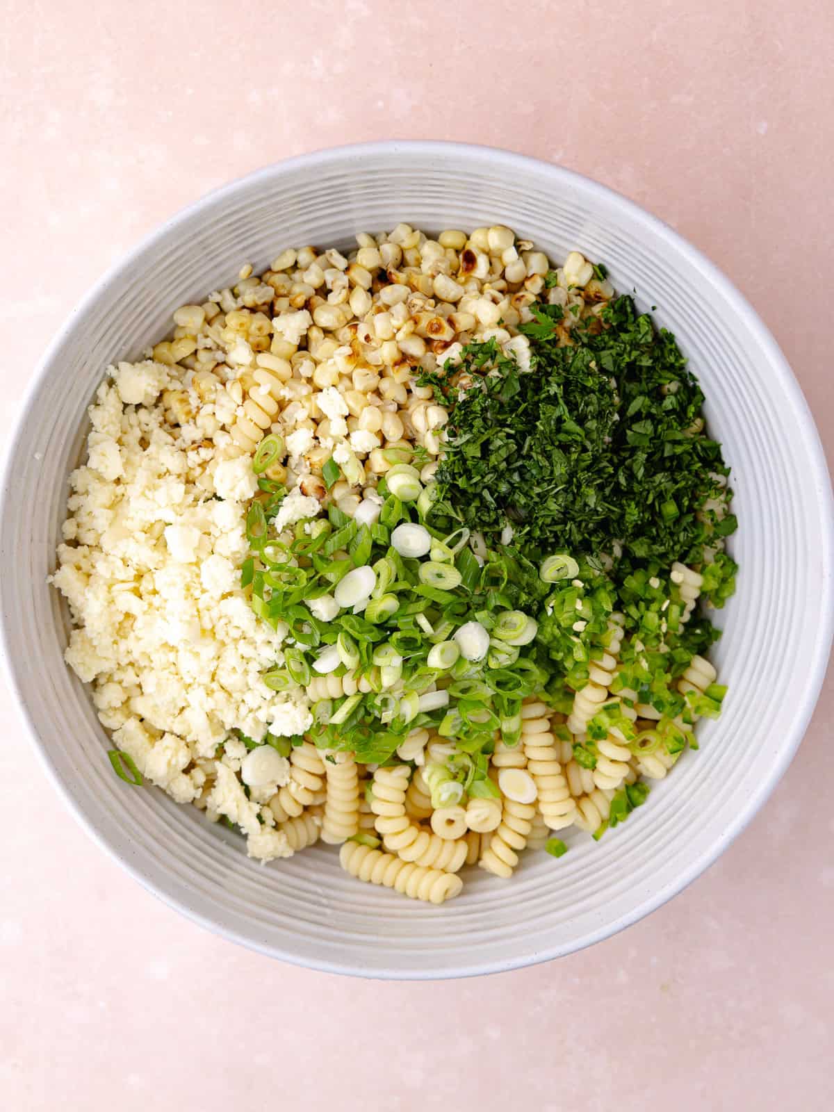 Cooked gemelli pasta, charred corn, crumbled cotija cheese, chopped green onions, finely chopped jalapeño, finely chopped cilantro are in a large bowl.