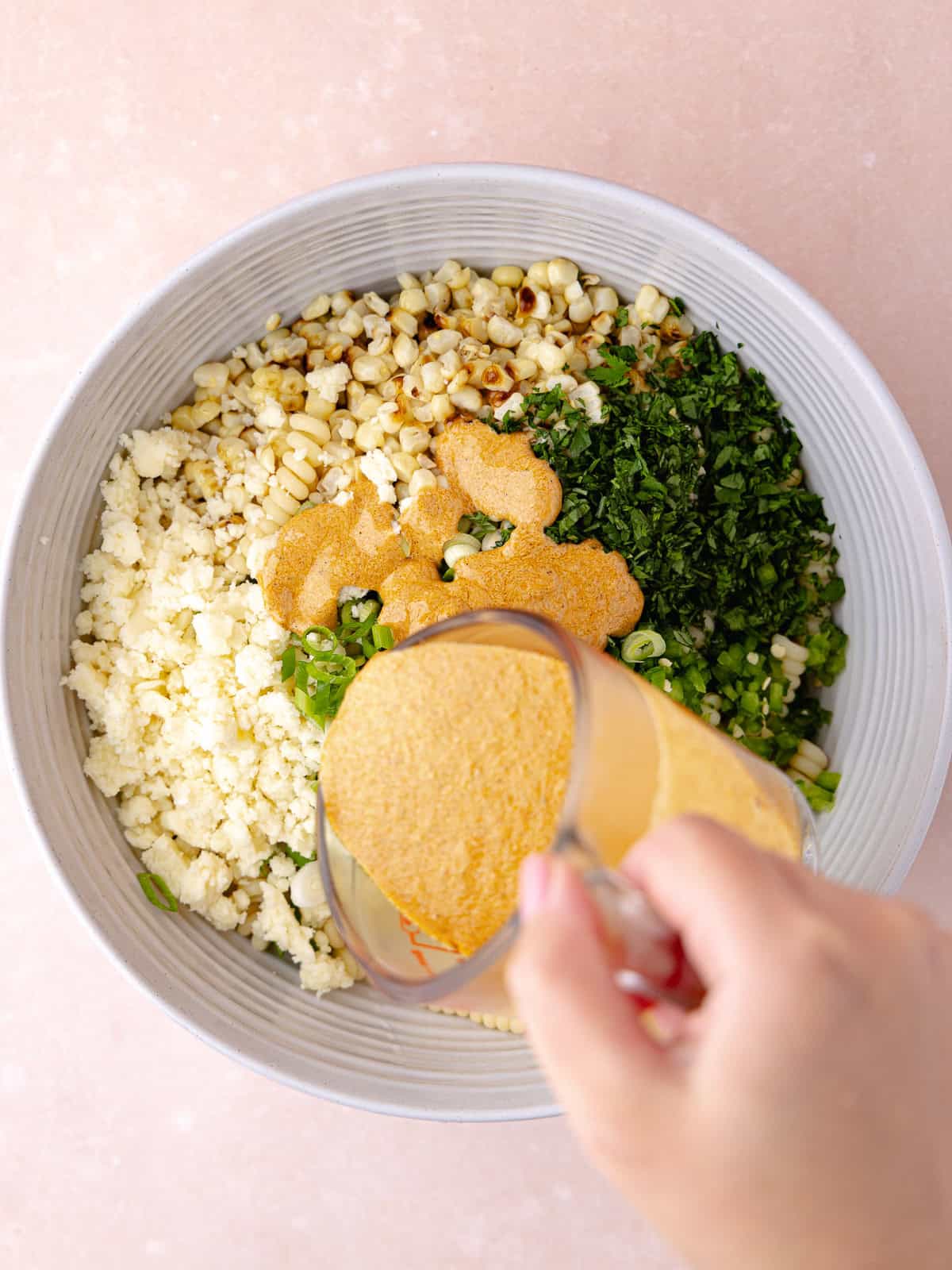 Creamy smokey Greek yogurt dressing is poured over the cooked gemelli pasta, charred corn, crumbled cotija cheese, chopped green onions, finely chopped jalapeño, finely chopped cilantro in a large bowl.