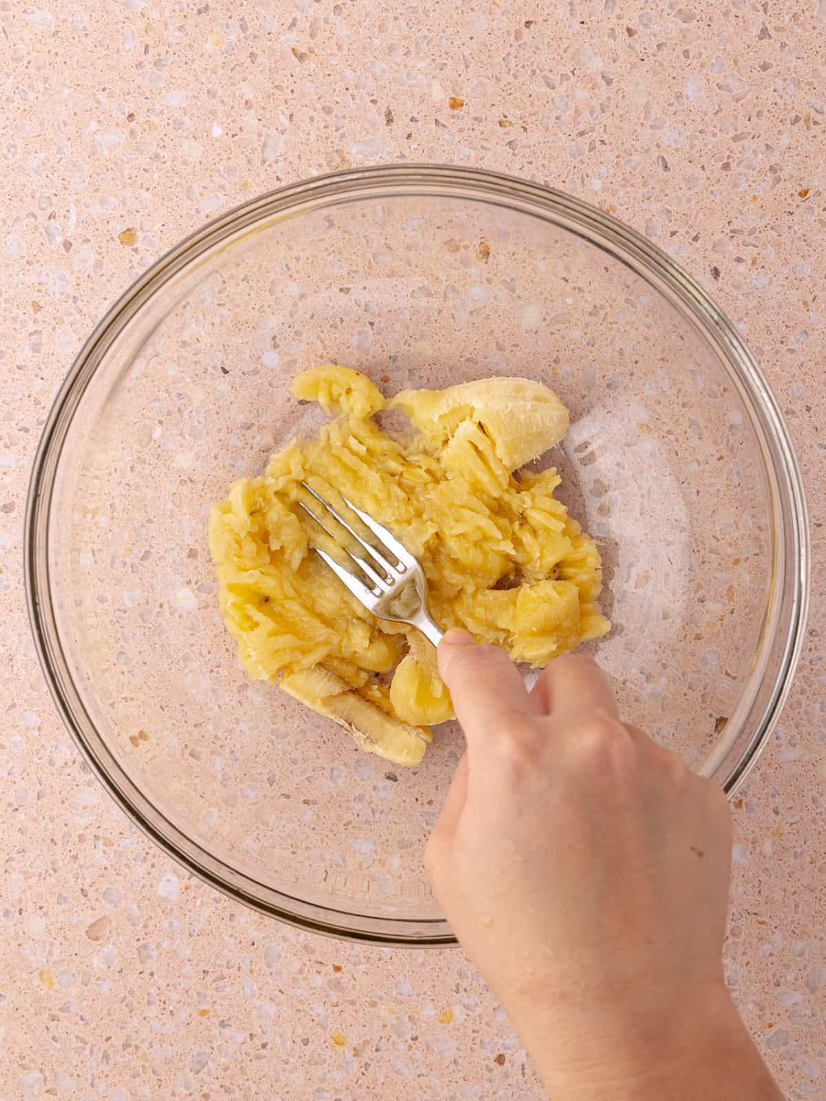 A ripe banana sitting in a glass mixing bowl is smashed with a fork.