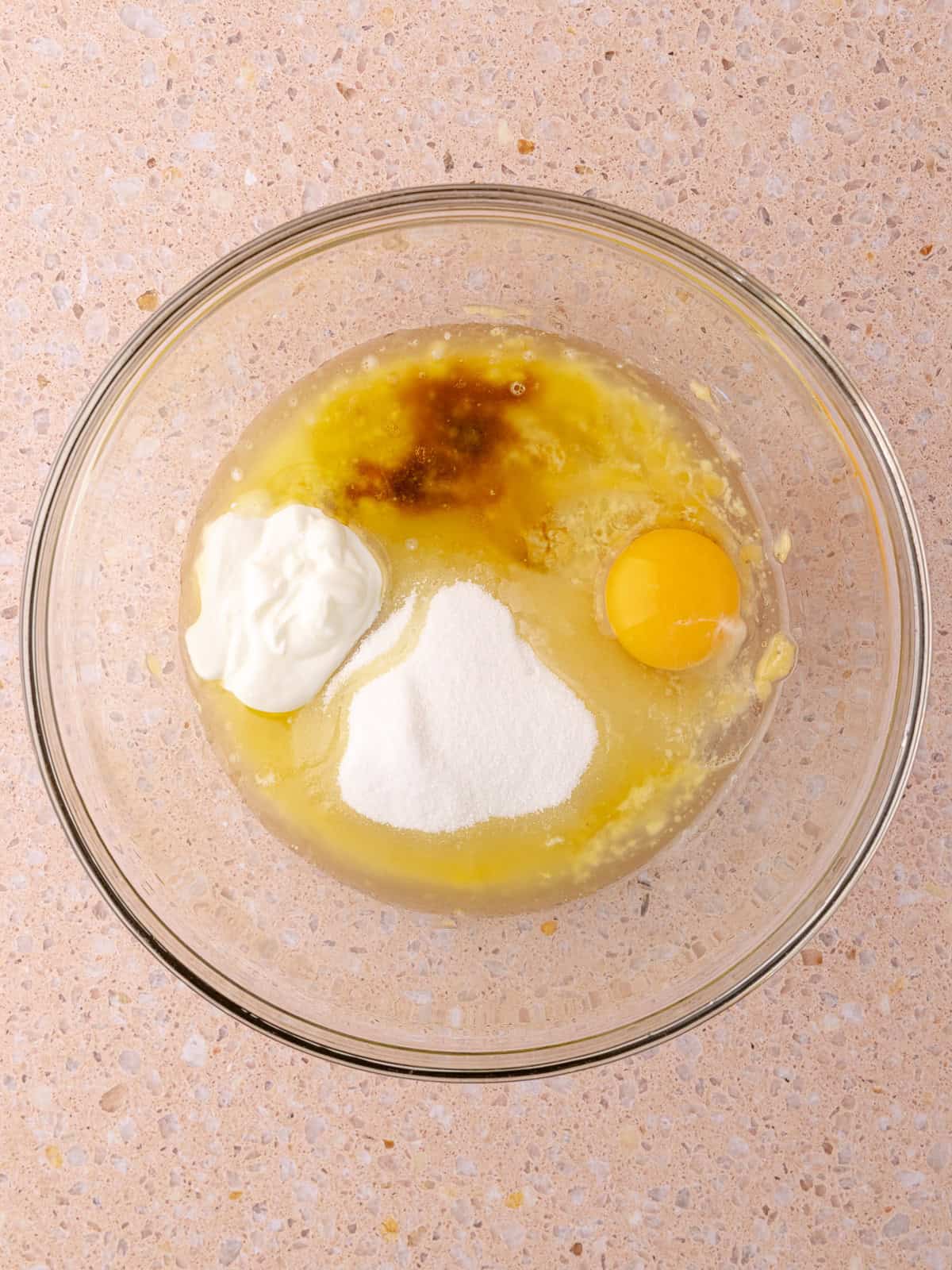 Mashed banana, melted butter, oil, sugar, Greek yogurt, vanilla extract and an egg are in a glass mixing bowl.