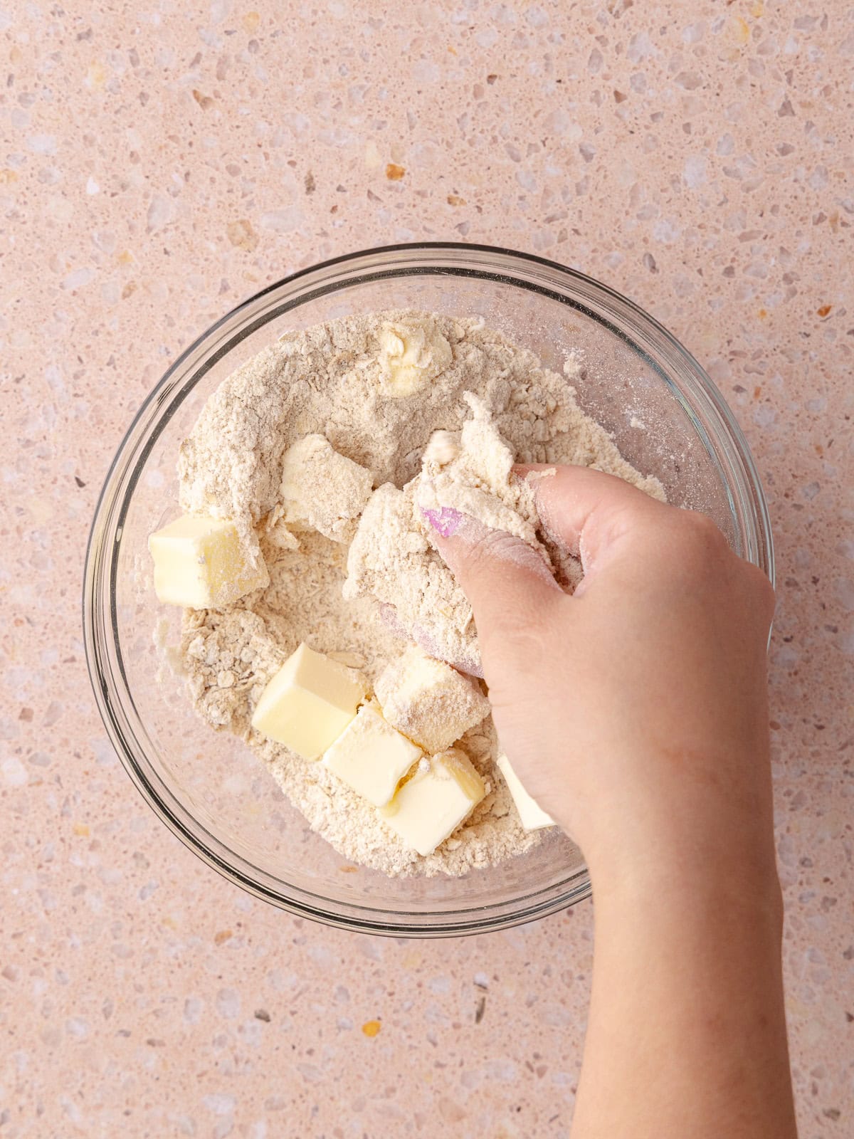 A person's hand combining flour, oats, brown sugar and butter in a small mixing bowl.