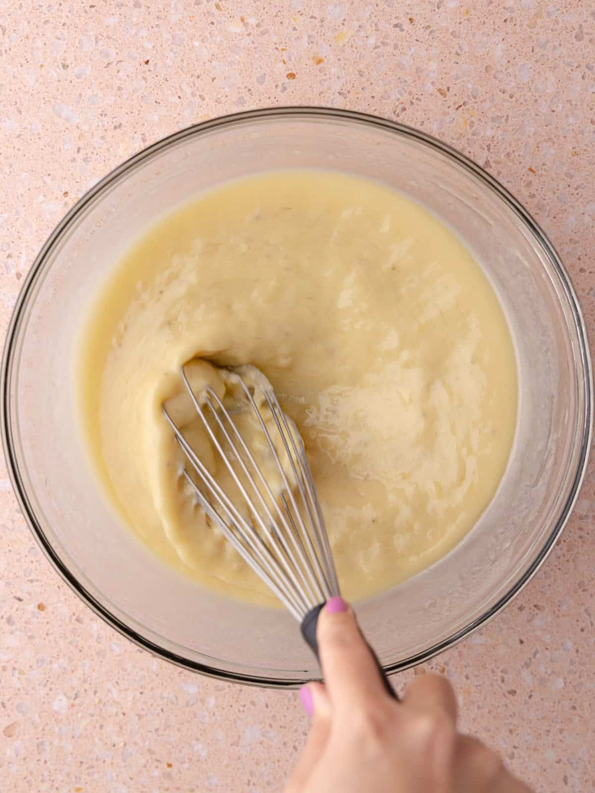 A person whisking wet ingredients in a large glass bowl.