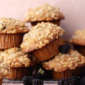A pile of banana blackberry oatmeal muffins with oatmeal streusel stacked on a wire rack with fresh blackberries feature photo.