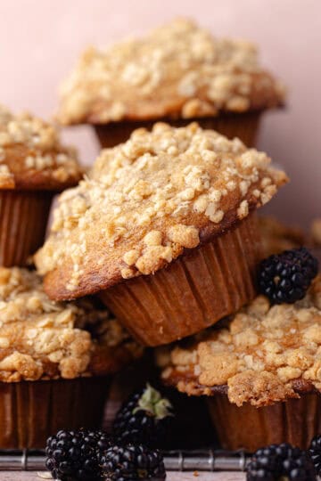 A pile of banana blackberry oatmeal muffins with oatmeal streusel stacked on a wire rack with fresh blackberries feature photo.