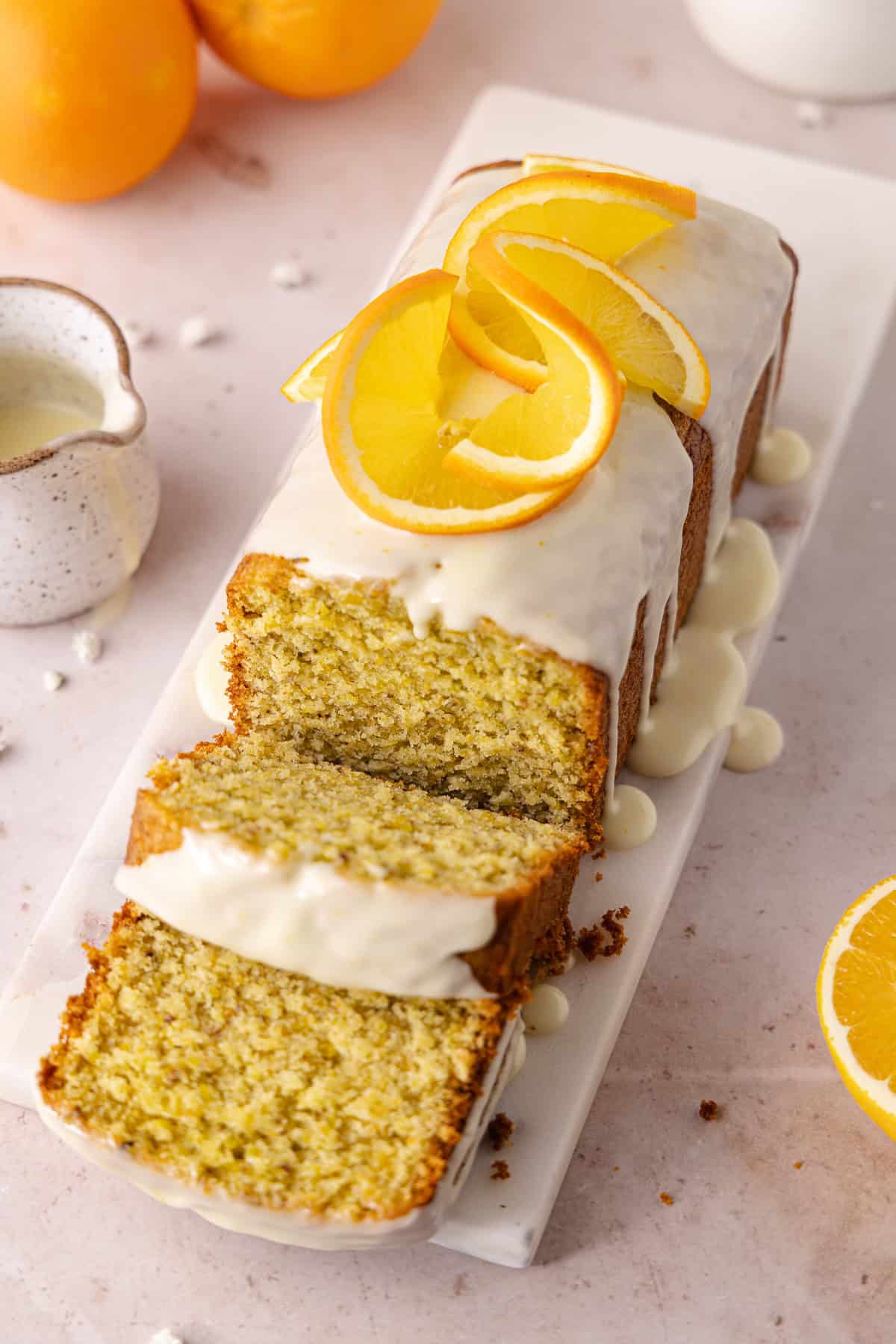 Pistachio Orange Olive Oil Cake topped with an orange glaze, is made with a whole orange and extra-virgin olive oil. Slices of Pistachio Orange Olive Oil Cake is sitting on a serving plate.