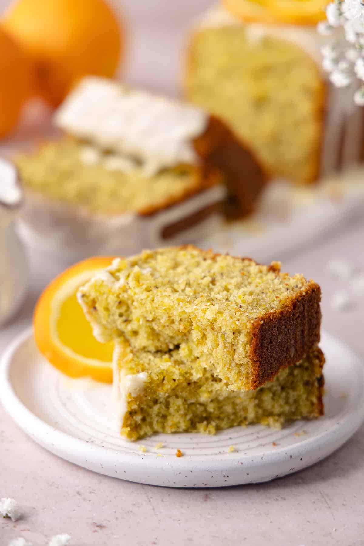 Pistachio Orange Olive Oil Cake topped with an orange glaze, is made with a whole orange and extra-virgin olive oil. A slice of Pistachio Orange Olive Oil Cake is on a small dessert plate.