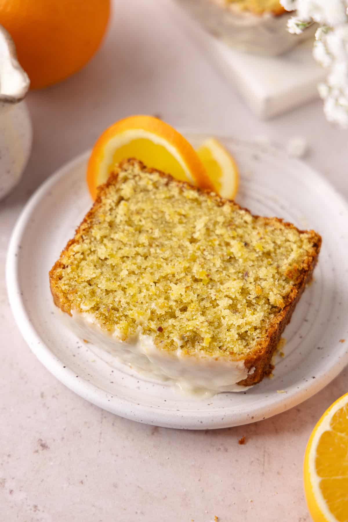 Pistachio Orange Olive Oil Cake topped with an orange glaze, is made with a whole orange and extra-virgin olive oil. A slice of Pistachio Orange Olive Oil Cake is on a small dessert plate.