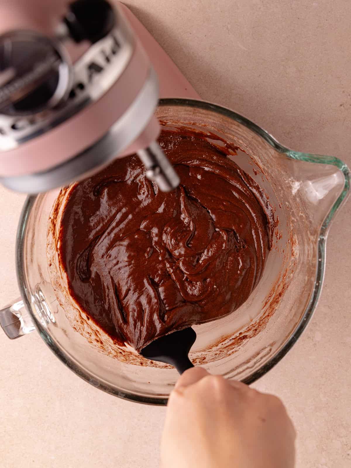A hand holding a silicone spatula scrapes down the bowl with chocolate cake batter in a stand mixer bowl.
