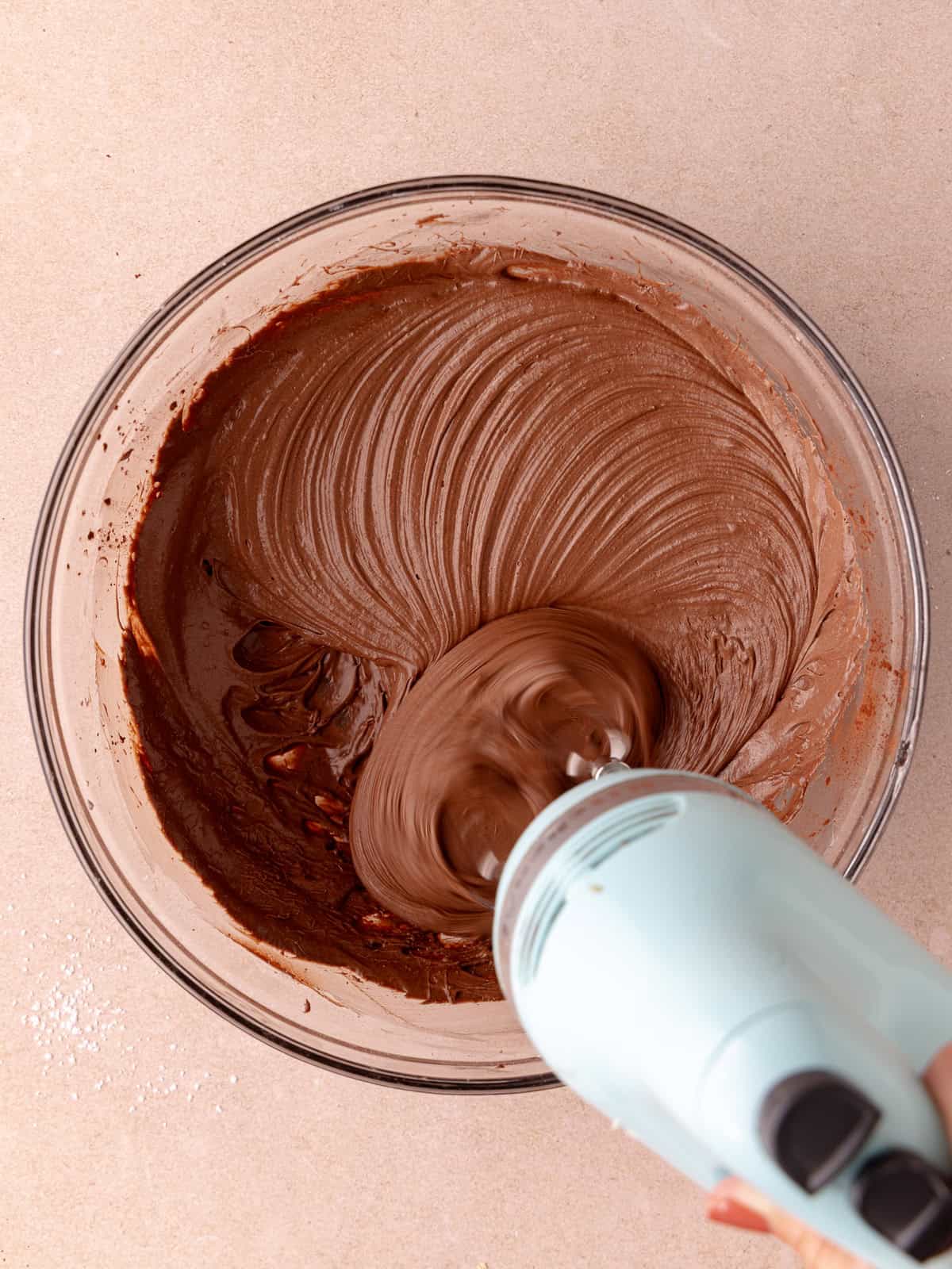 Chocolate malted frosting is whipped with an electric mixer in a large mixing bowl.