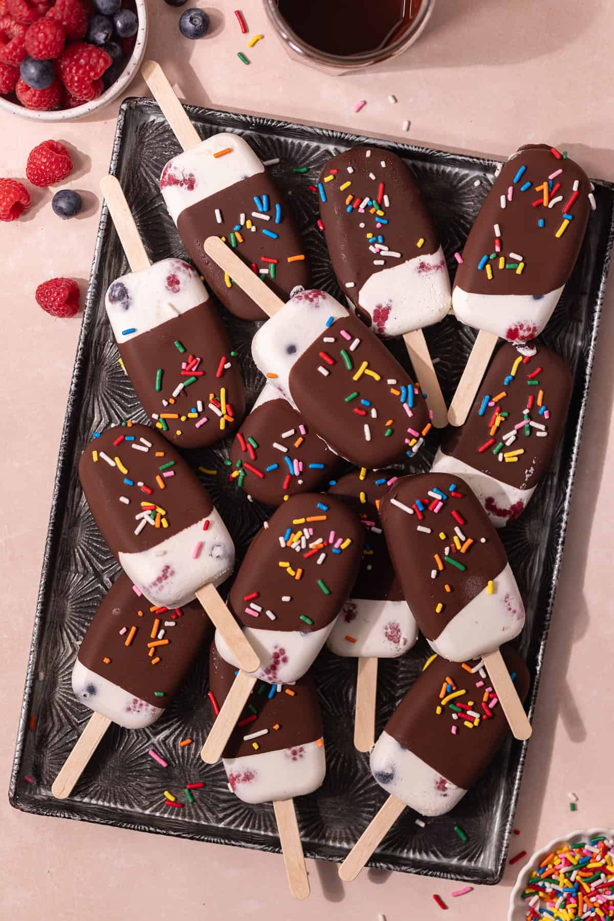 Creamy coconut popsicles with fresh raspberries and blueberries are dipped in a magic chocolate shell and topped with rainbow sprinkles.