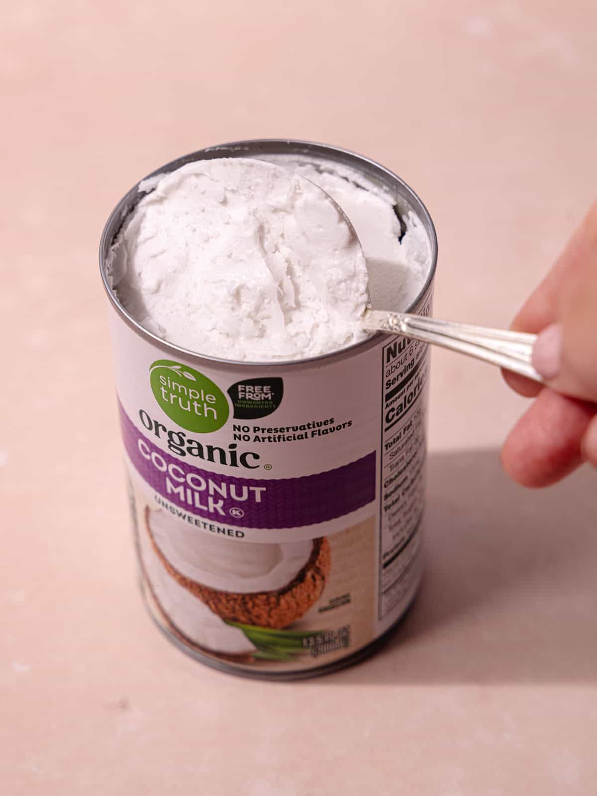Canned coconut milk is refrigerated to separate the coconut cream layer from the coconut water. A hand scoops the firm layer of coconut cream with a spoon.
