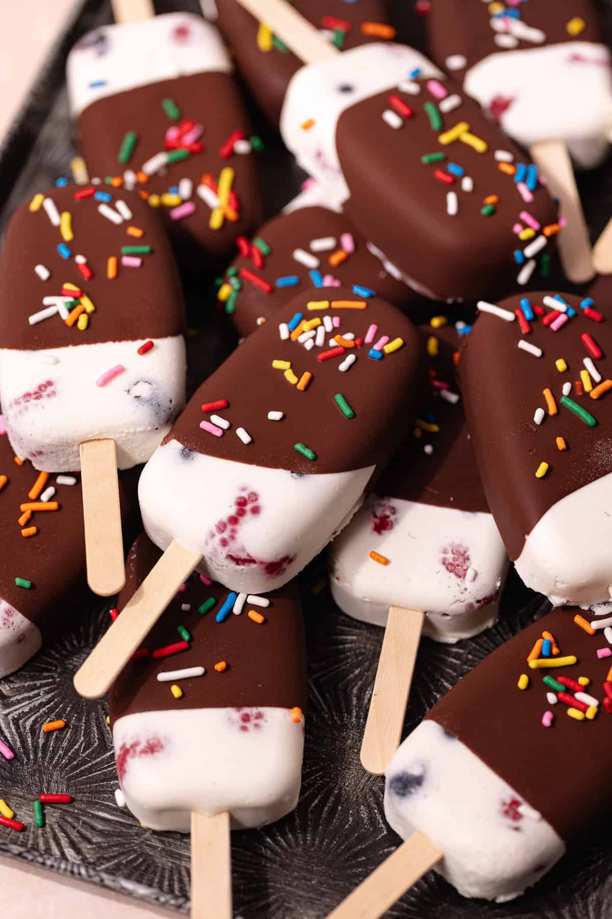 Creamy coconut popsicles with fresh raspberries and blueberries are dipped in a magic chocolate shell and topped with rainbow sprinkles.