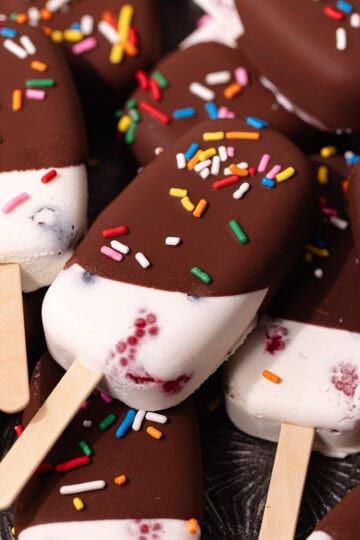 Creamy coconut popsicles with fresh raspberries and blueberries are dipped in a magic chocolate shell and topped with rainbow sprinkles.