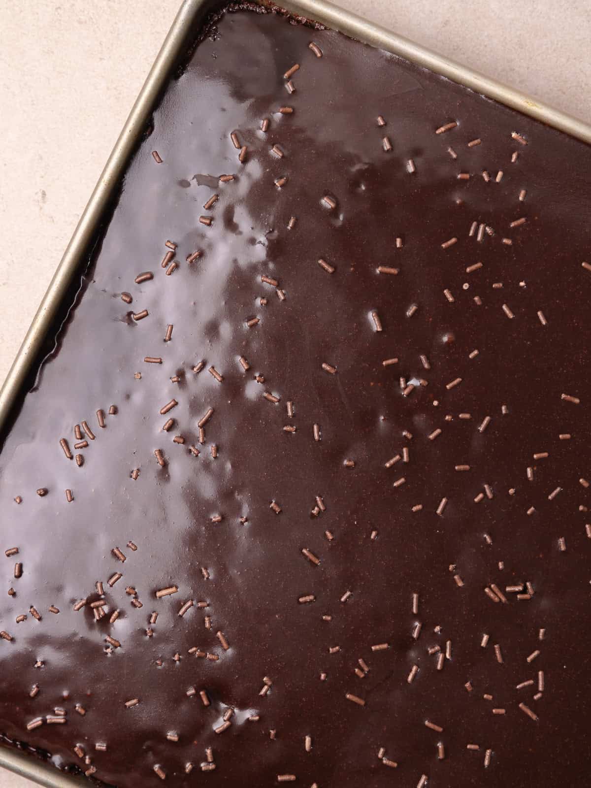 The warm chocolate icing is poured over the chocolate sheet cake and spread to cover the top of the cake. It's then garnished with chocolate sprinkles.
