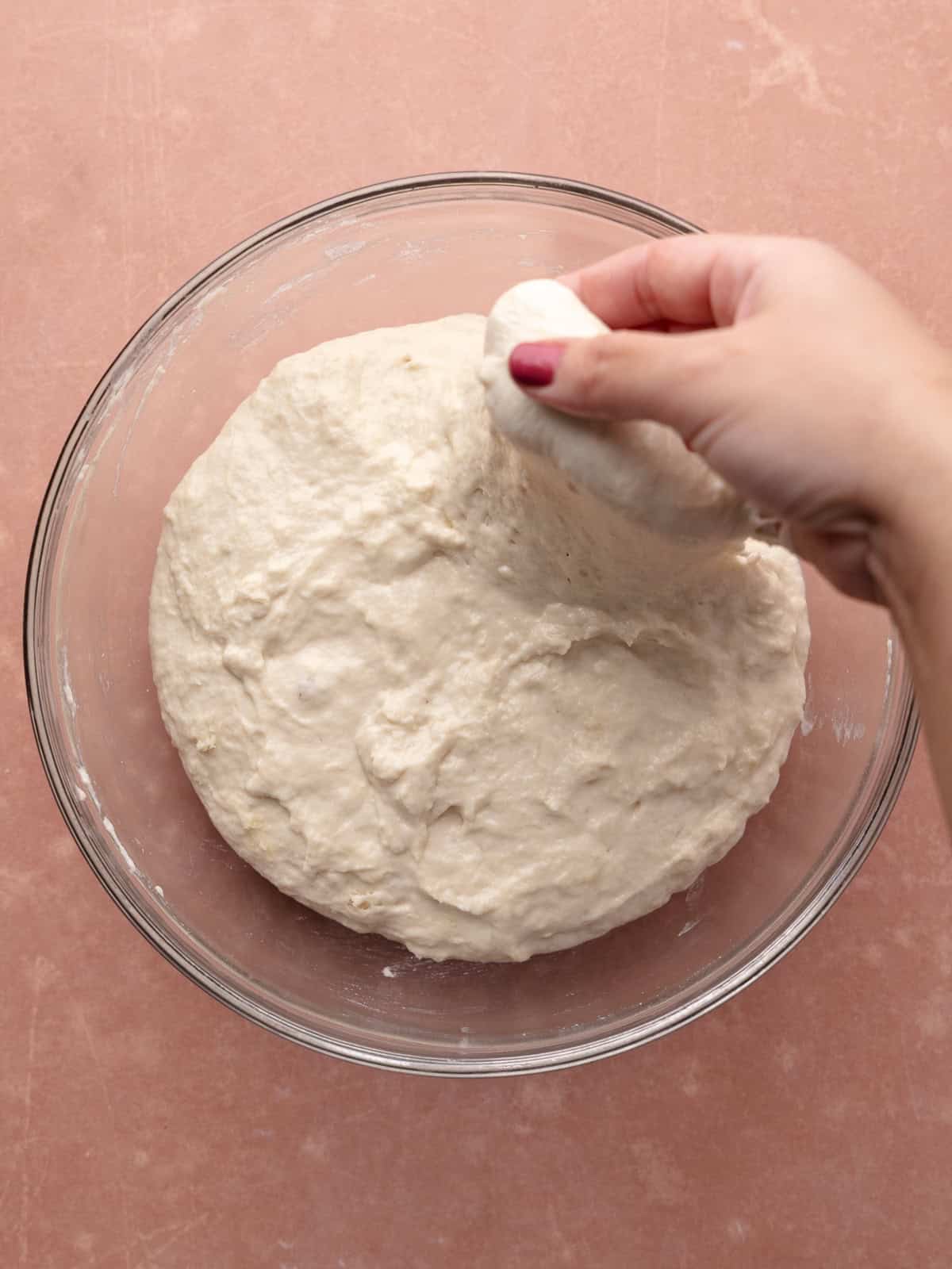A hand picks up a side of the focaccia dough and stretches it upwards.