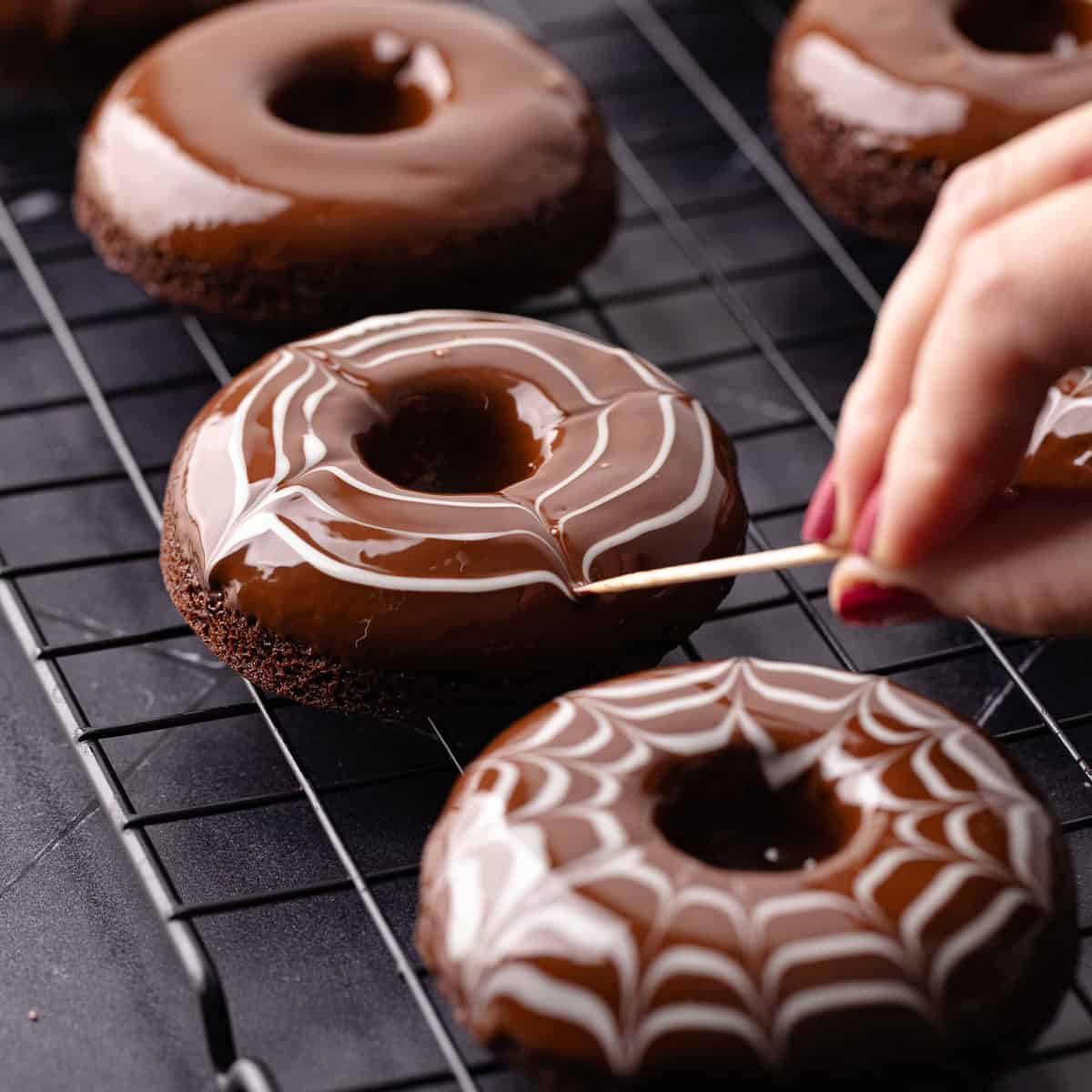 Baked chocolate donuts are dipped in semi-sweet chocolate and then white chocolate is dragged on top of the chocolate to create a spider web effect.