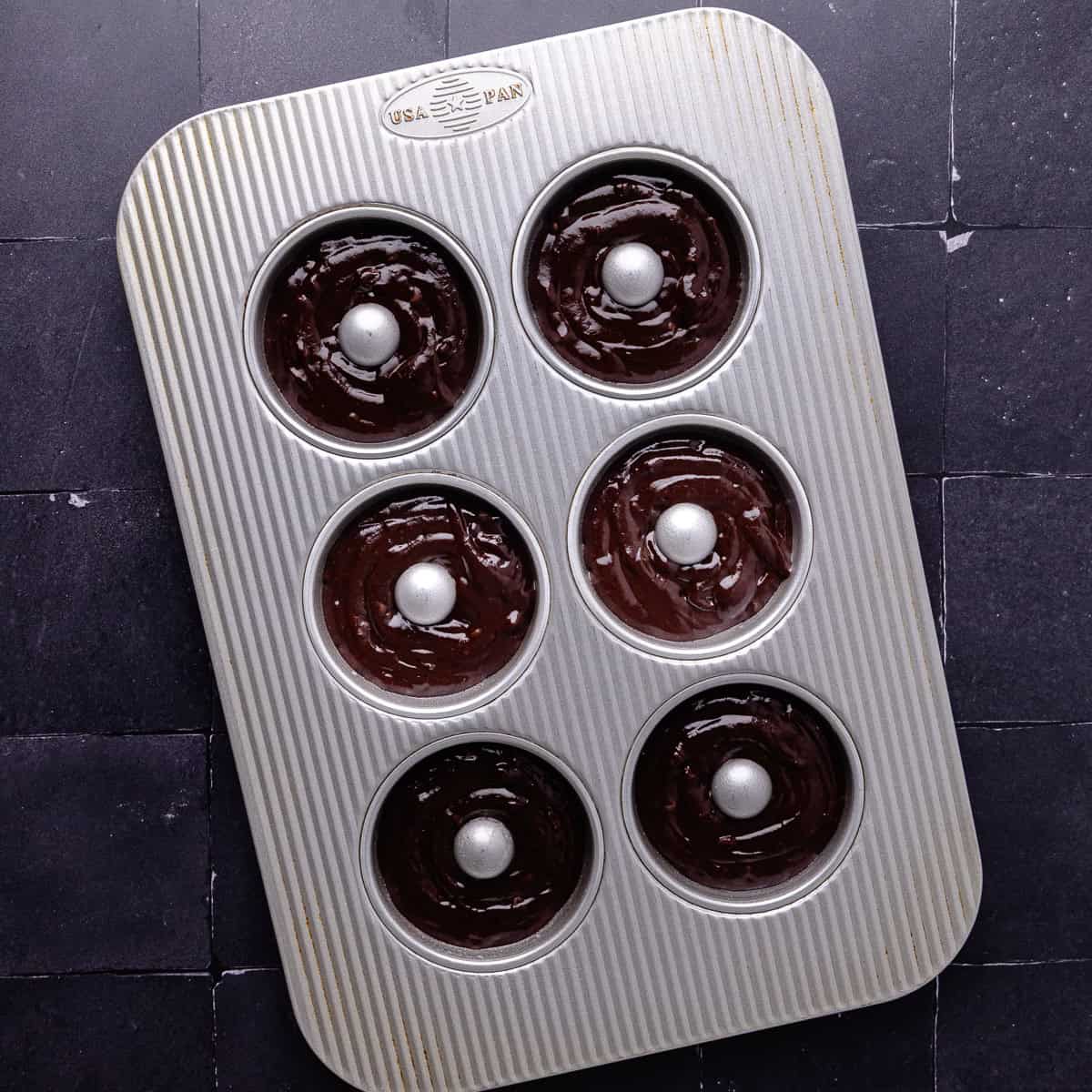 Chocolate donut batter with mini chocolate chips are piped into a 6-cavity donut pan.