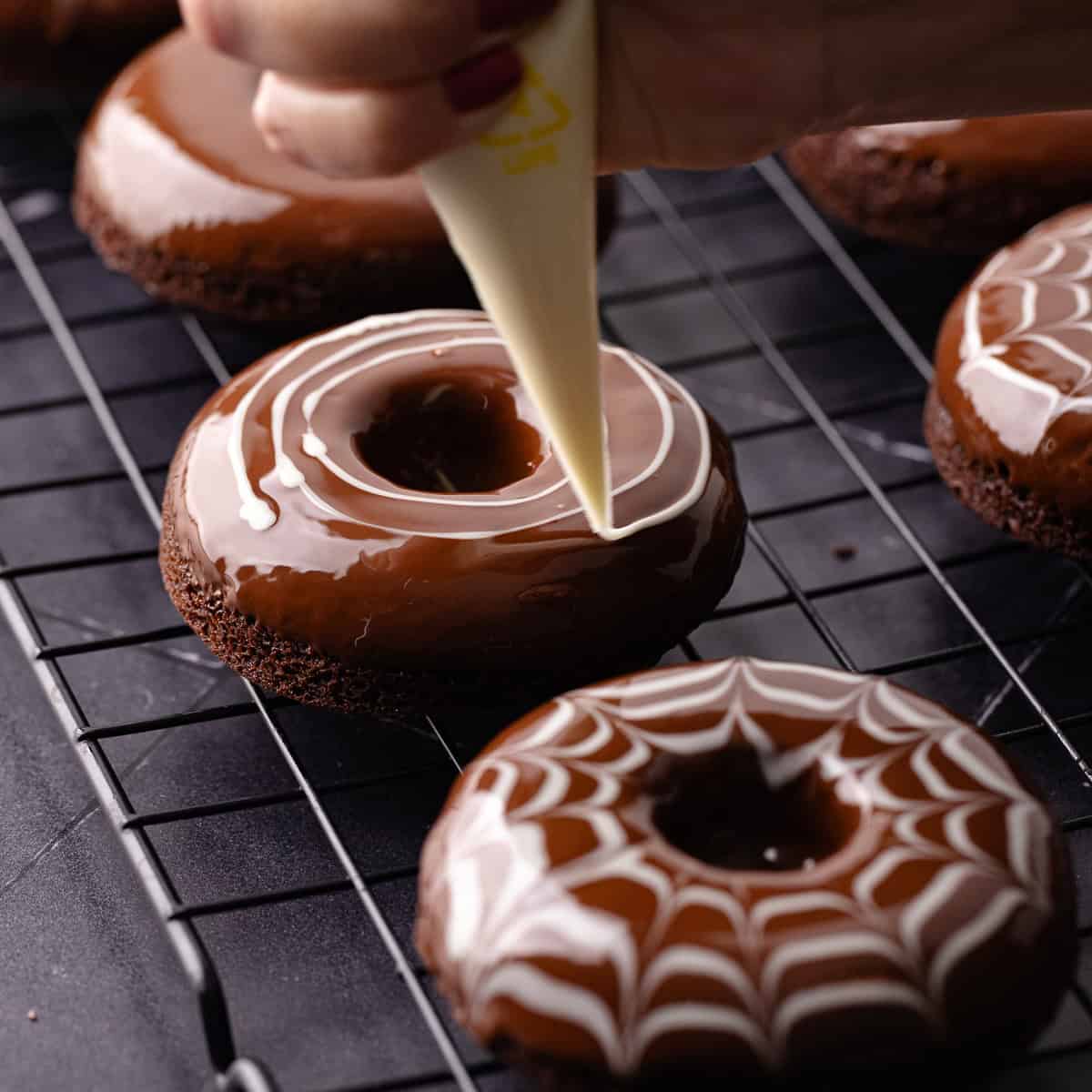 Baked chocolate donuts are dipped in semi-sweet chocolate and then white chocolate is dragged on top of the chocolate to create a spider web effect.